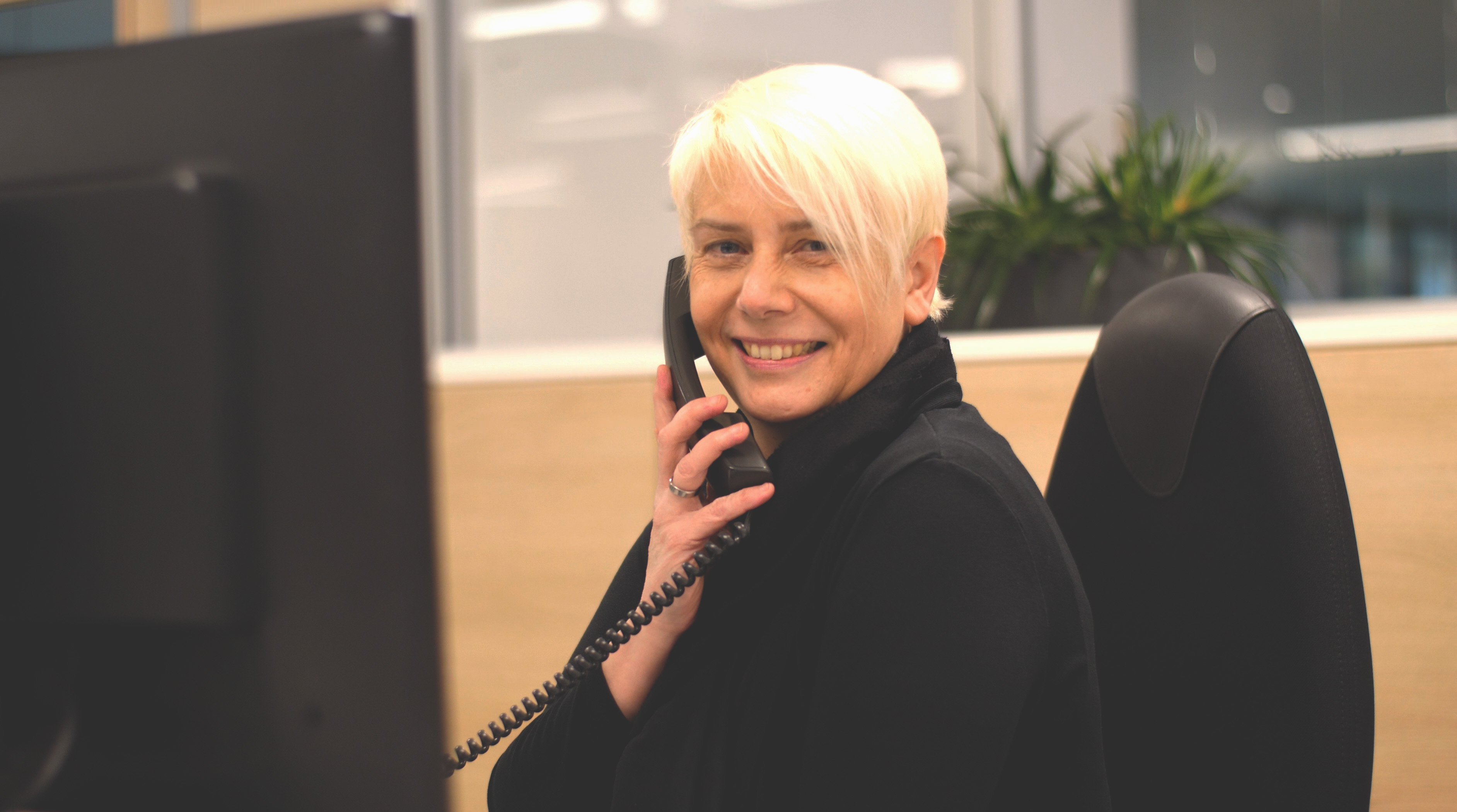 Eine lächelnde Frau mit kurzen, blonden Haaren telefoniert an einem Schreibtisch in einem Büro.